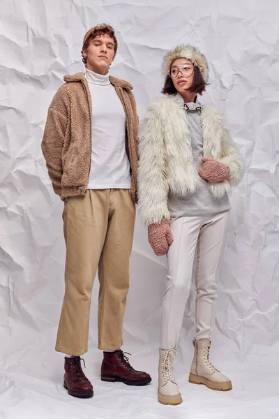 Hombre de moda mirando a la cámara cerca de mujer asiática en chaqueta caliente y manoplas sobre fondo texturizado blanco - foto de stock