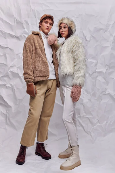 Asian woman in faux fur jacket and hat leaning on shoulder of trendy man on white textured backdrop — Stock Photo