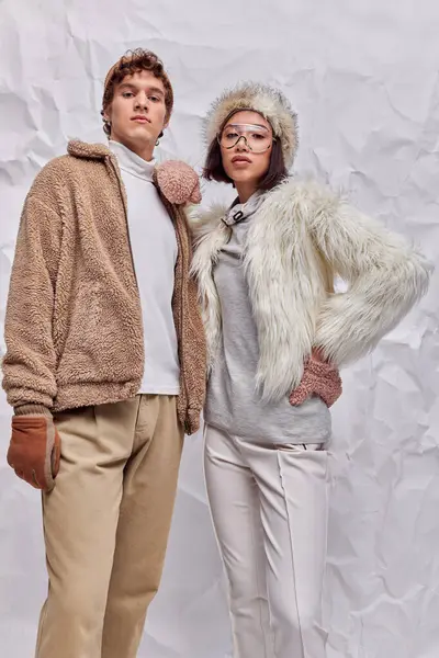 Trendy asian woman in warm clothes posing with hand on hip near young man on white textured backdrop — Stock Photo