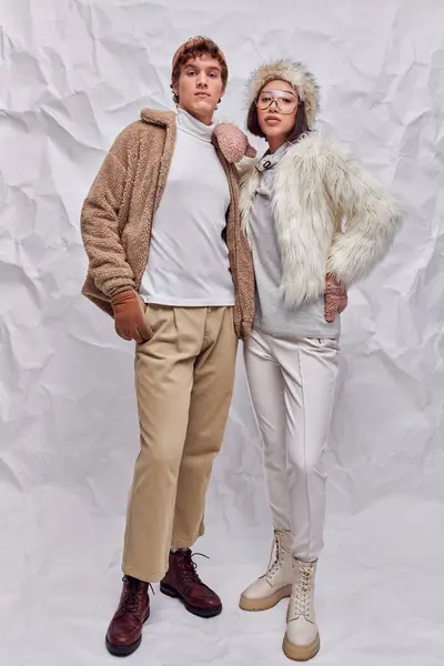 Young asian model in warm attire posing with hand on hip near trendy man on white textured backdrop — Stock Photo