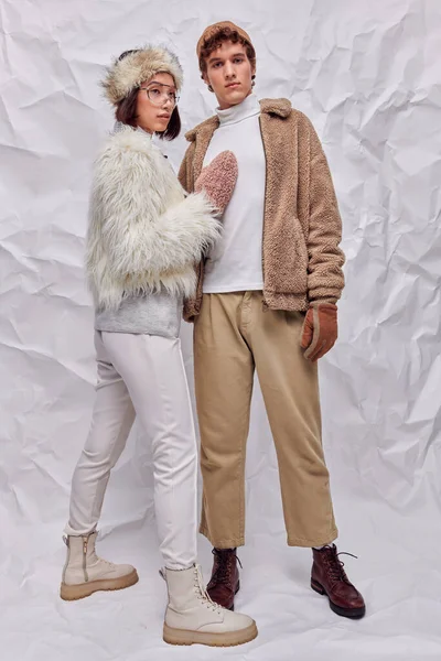 Jeune homme à la mode regardant caméra près de femme asiatique dans des vêtements chauds et confortables sur fond blanc froissé — Photo de stock
