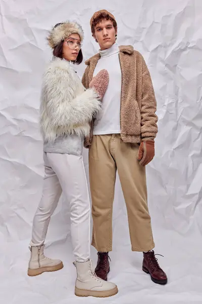 Young trendy man looking at camera near asian woman in warm cozy clothes on white crumpled backdrop — Stock Photo