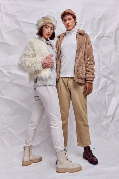 Концепция модной кампании, межрасовая пара в искусственных меховых куртках позирует на белом скомканном фоне — стоковое фото