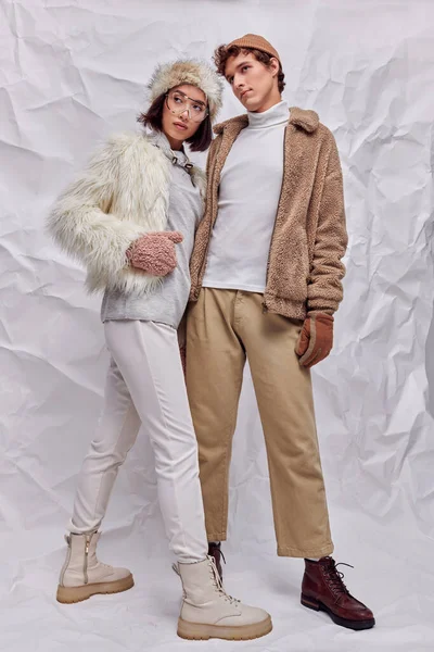 Многонациональная пара в модной зимней одежде, смотрящая в сторону на белом текстурированном фоне, сезонный стиль — стоковое фото