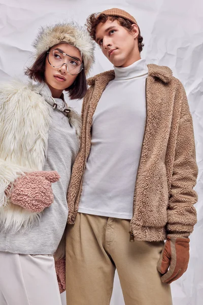 Mode d'hiver, couple interracial en vêtements d'extérieur chauds regardant loin sur fond texturé blanc — Photo de stock