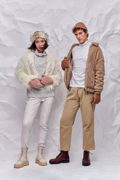 Interracial couple in hats, warm gloves and faux fur jackets standing on white crumpled backdrop — Stock Photo