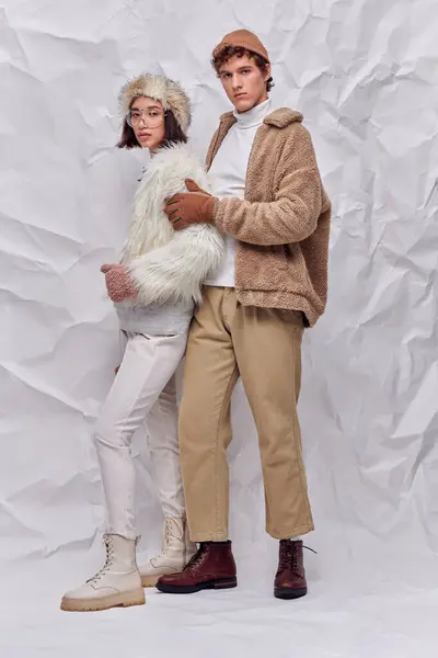 Young stylish man embracing asian woman in faux fur jacket and eyeglasses on crumpled paper backdrop — Stock Photo