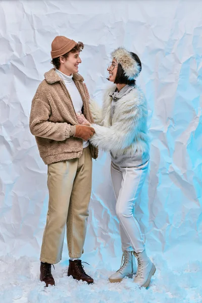 Feliz pareja interracial en ropa de invierno de moda mirándose el uno al otro en la nieve y fondo blanco - foto de stock