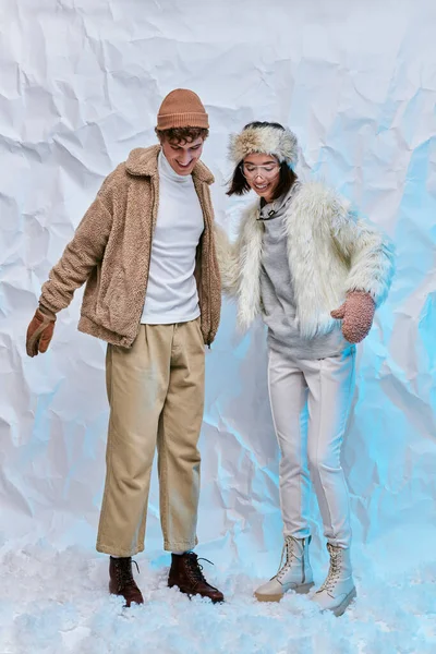 Joyful interracial couple in trendy winter clothes standing on snow on white textured backdrop — Stock Photo