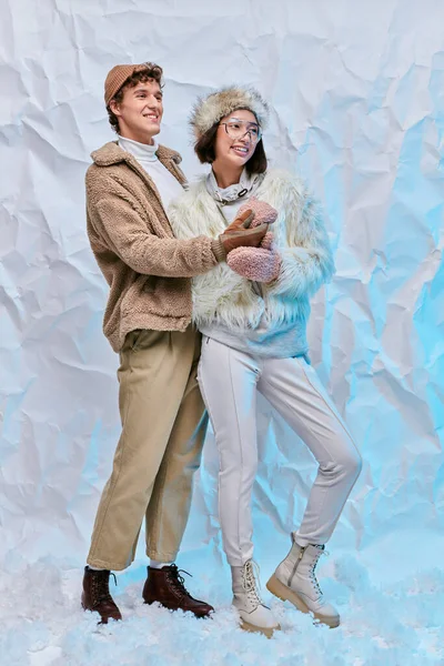 Беззаботная межрасовая пара в стильной зимней одежде, смотрящая на снег на белом текстурированном фоне — стоковое фото