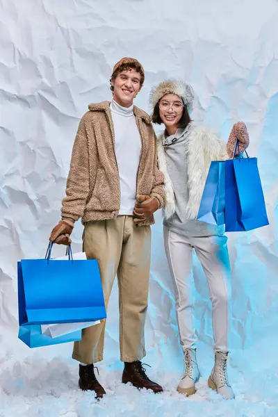 Joyful multiethnic models in warm outerwear with blue paper bags on snow in studio, winter shopping — Stock Photo