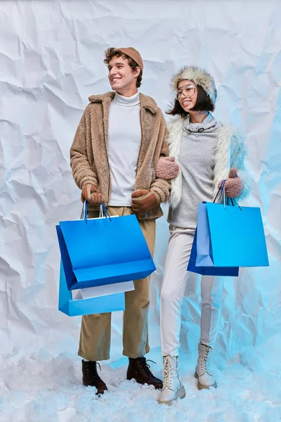 Щаслива міжрасова пара в зимовому одязі з блакитними сумками в засніженій студії, сезонні покупки — стокове фото