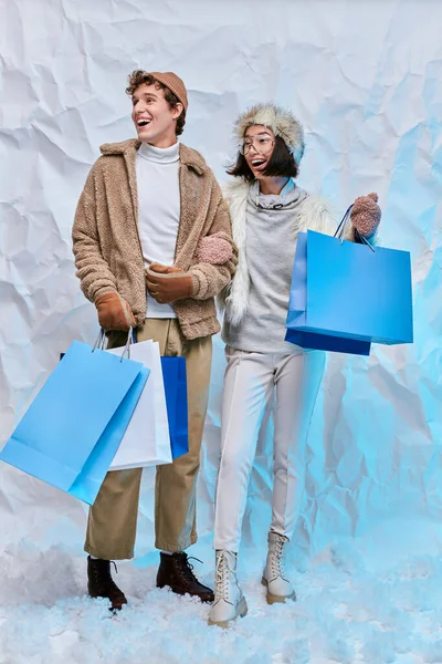 Amazed interracial couple in winter attire with shopping bags looking away on white snow in studio — Stock Photo