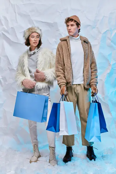 Winter shopping, multiethnic couple in seasonal clothes with white shopping bags on snow in studio — Stock Photo