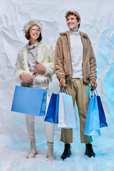 Зимняя модная кампания, счастливая межрасовая пара с голубыми сумками на белом снегу в студии — стоковое фото