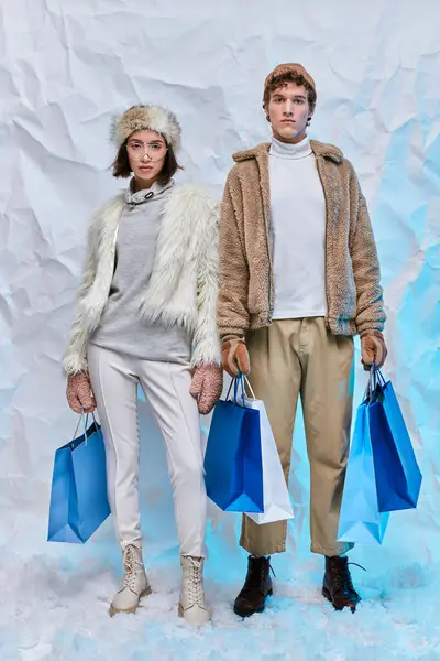 Trendy interracial models in winter wear with blue shopping bags looking at camera in snowy studio — Stock Photo