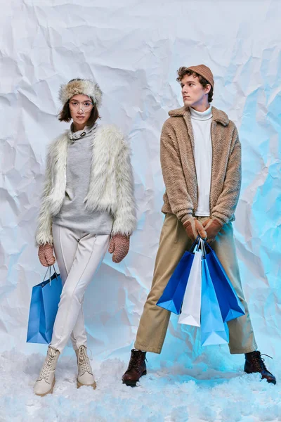 Модная межрасовая пара позирует с голубыми сумками для покупок на белом снегу в студии, зимняя мода — стоковое фото