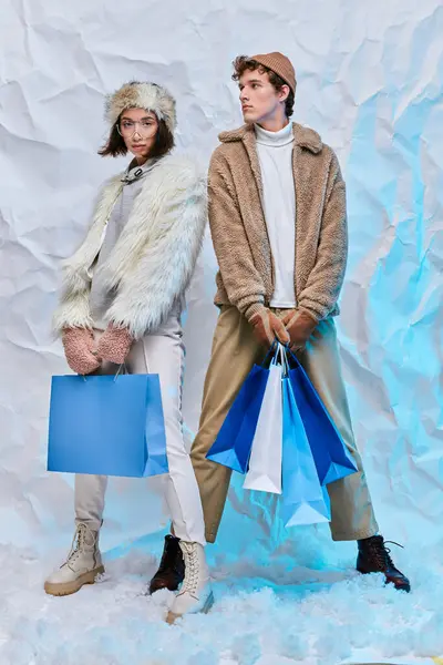 Shopping d'hiver, modèles de mode interracial dans une tenue chaleureuse et confortable avec des sacs à provisions dans un studio enneigé — Photo de stock