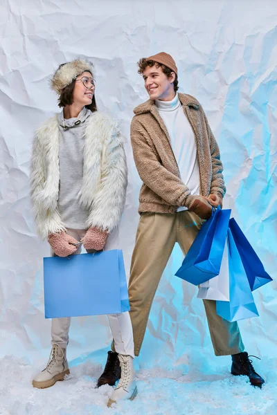 Модна міжрасова пара з сумками, посміхаючись один одному на снігу в студії, зимовий стиль — стокове фото