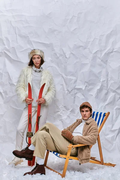 Зимова мода і дозвілля, азіатська жінка з лижами біля модного чоловіка в кріслі на палубі в засніженій студії — стокове фото