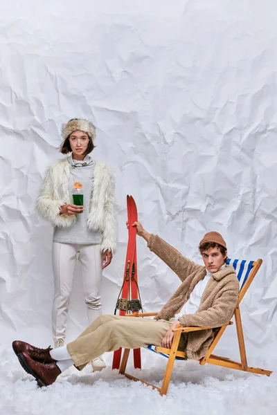 Stylish man with skis sitting in deck chair near asian woman with hot cocktail on snow in studio — Stock Photo