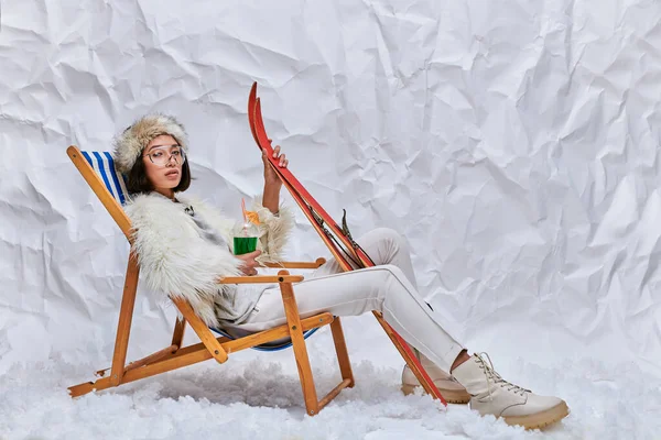 Asiatin in Winterbekleidung sitzt im Liegestuhl mit Skiern und heißem Teddycocktail auf Schnee im Studio — Stockfoto