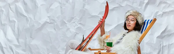 Donna asiatica in abbigliamento invernale in sedia a sdraio con sci e cocktail su sfondo bianco strutturato, banner — Foto stock