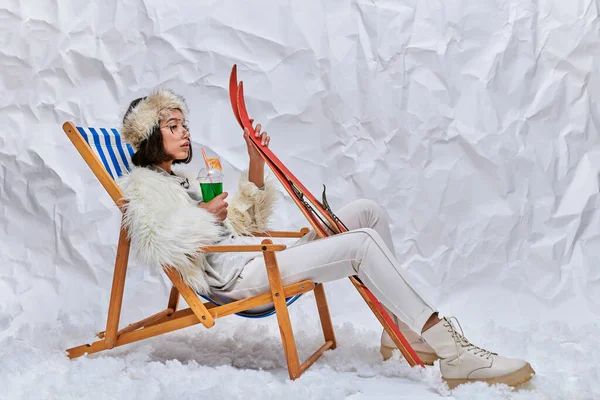Asian woman in warm trendy outfit with skis and cocktail relaxing in deck chair on snow in studio — Stock Photo