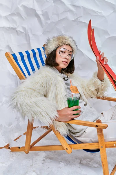 Glamour asiatique femme en fausse fourrure veste en chaise longue avec cocktail et skis sur fond blanc — Photo de stock