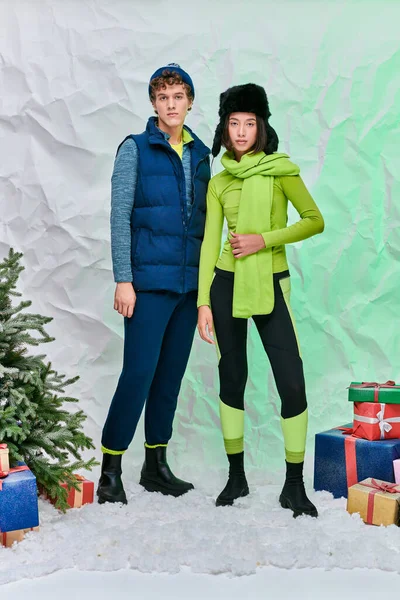 Pareja interracial en ropa de invierno brillante cerca de cajas de regalo y árbol de Navidad en la nieve en el estudio - foto de stock