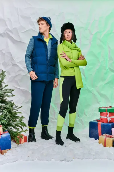 Pareja multiétnica en ropa caliente cerca de cajas de regalo y árbol de Navidad en la nieve en el estudio, temporada festiva - foto de stock