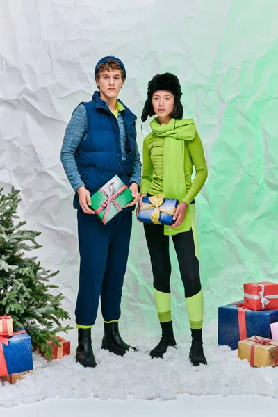 Модная межрасовая пара в зимней одежде с подарками возле рождественской елки на снегу в студии — стоковое фото