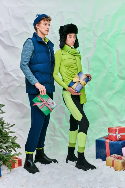 Elegante casal interracial com caixas de presente olhando para longe perto da árvore de natal no estúdio nevado — Fotografia de Stock