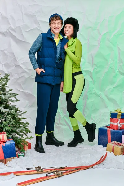 Happy interracial fashion models in winter outfit near christmas tree and presents on snow in studio — Stock Photo