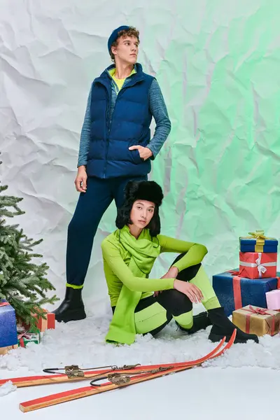Asian model sitting on snow in studio near stylish man, skis and gift boxes with christmas tree — Stock Photo