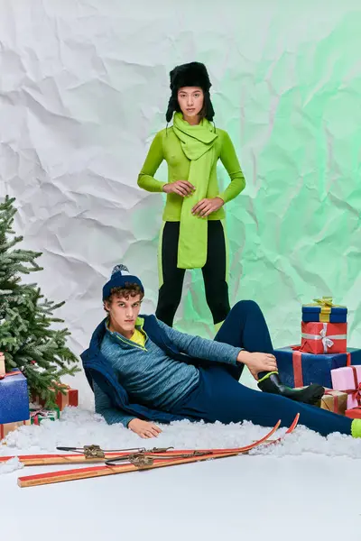 Asiatin blickt in die Kamera neben trendigem Mann auf Schnee in der Nähe von Geschenken und Weihnachtsbaum im Atelier — Stockfoto