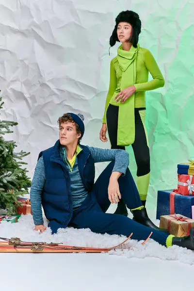 Coppia interrazziale in inverno indossare guardando lontano accanto ai regali e albero di Natale in studio innevato — Foto stock