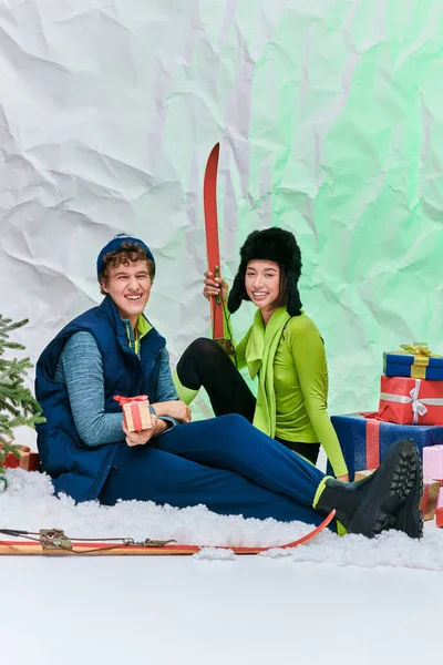 Alegre asiático modelo con esquís sentado cerca de elegante hombre, árbol de Navidad y regalos en nevado estudio - foto de stock