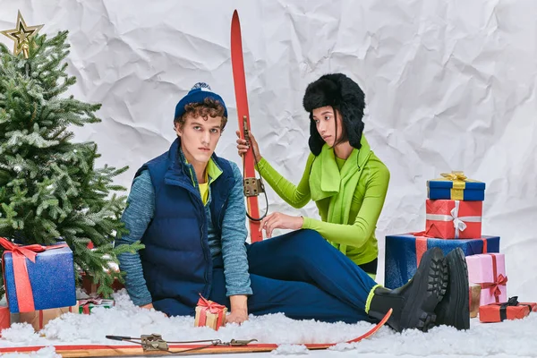 Молодая азиатская модель с лыжами сидит на снегу в студии возле модного человека, елка и подарочные коробки — стоковое фото