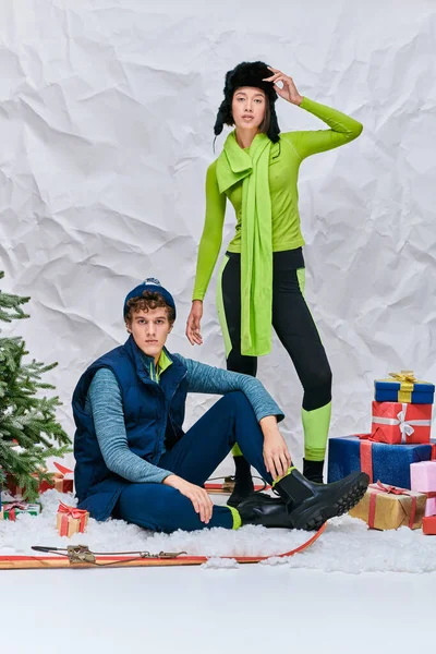 Яркая межрасовая пара, смотрящая в камеру возле подарков и рождественской елки в снежной студии — стоковое фото