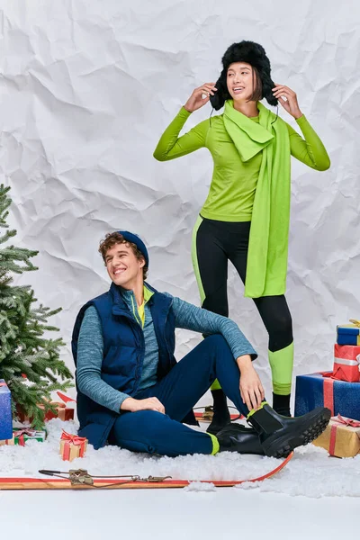 Веселая многонациональная пара, смотрящая в заснеженной студии возле рождественской елки и подарочных коробок — стоковое фото