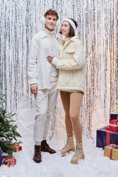 Happy asian woman looking at stylish man near presents and christmas tree on shiny tinsel backdrop — Stock Photo