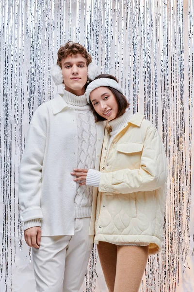 Joyful interracial couple in white jackets standing near silver tinsel on backdrop, christmas spirit — Stock Photo