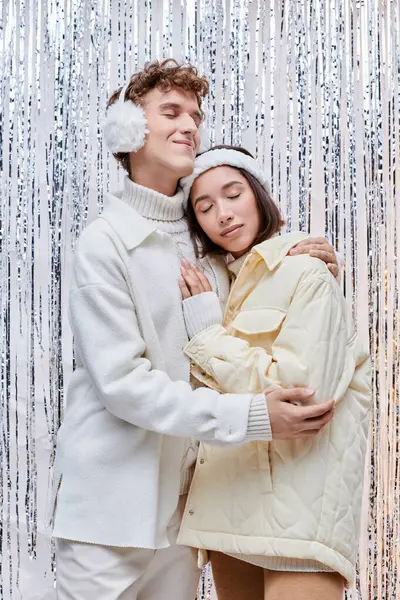 Happy interracial couple in cozy winter wear embracing with closed eyes near shiny tinsel in studio — Stock Photo