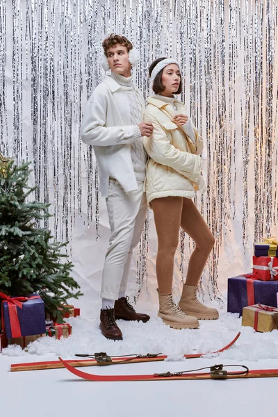 Межрасовая пара в зимнем наряде в студии возле серебряной мишуры и рождественских подарков на снегу — стоковое фото