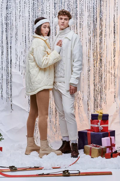 Couple interracial à la mode vêtements d'hiver en regardant la caméra près de cadeaux de Noël et argent tinsel — Photo de stock