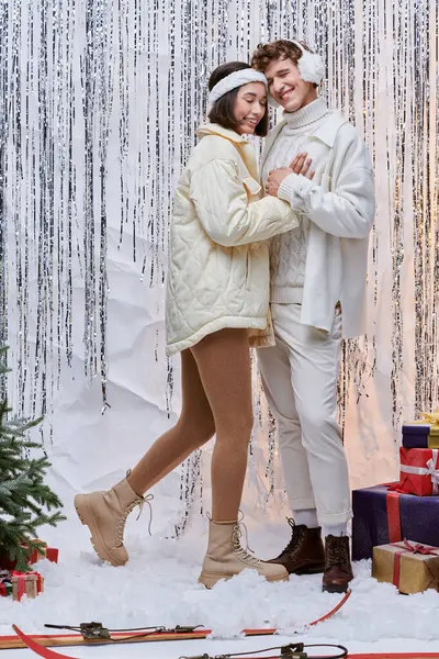 Молодая, стильная межрасовая пара улыбается рядом с подарками и рождественской елкой на блестящем фоне мишуры — стоковое фото