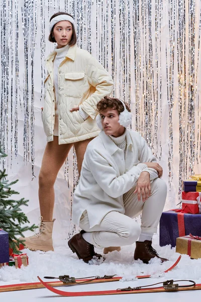 Modelos interracial en traje cálido posando cerca del árbol de Navidad, regalos y decoración de plata en el estudio - foto de stock