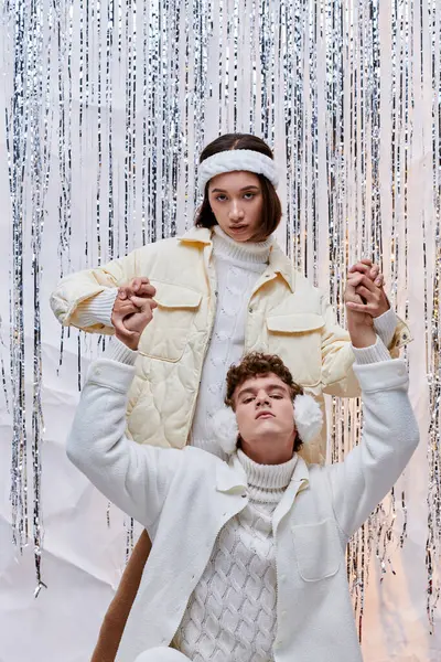 Pareja interracial de moda tomados de la mano mientras posan cerca de oropel brillante en el estudio, estilo de invierno - foto de stock