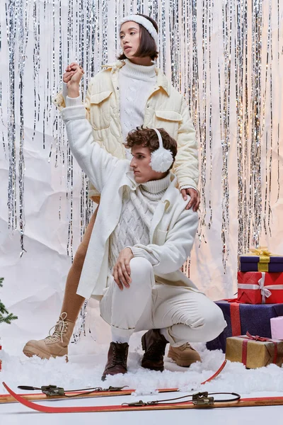Stylish asian woman holding hand of man in earmuffs posing near christmas gifts and silver tinsel — Stock Photo